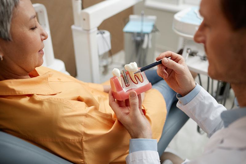 Closeup of male dentist holding tooth model while explaining dental implant surgery to a new patient in a dental clinic.